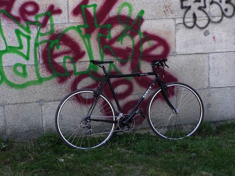 Right side view of a black Surly bike, leaning against a stone block wall with graffiti painted on it