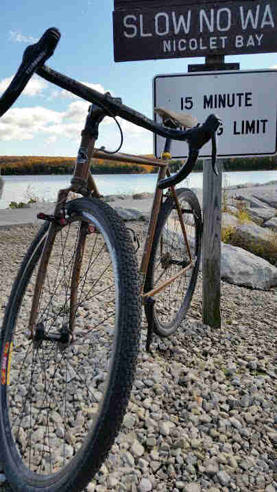Front view of a Surly bike, parked on a gravel bank in front of a sign, with a lake and trees in the background