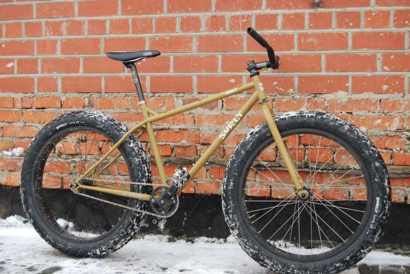 Right side view of a mustard yellow Surly Pugsley fat bike, parked on snowy ground, leaning against a brick wall