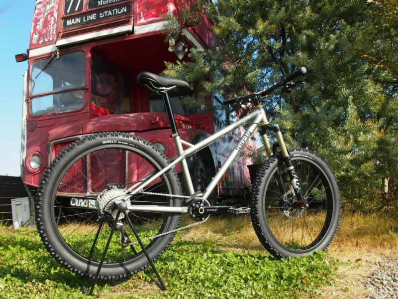 Right side view of a blue Surly Instigator bike, parked on grass, with a red double decker bus and a tree behind