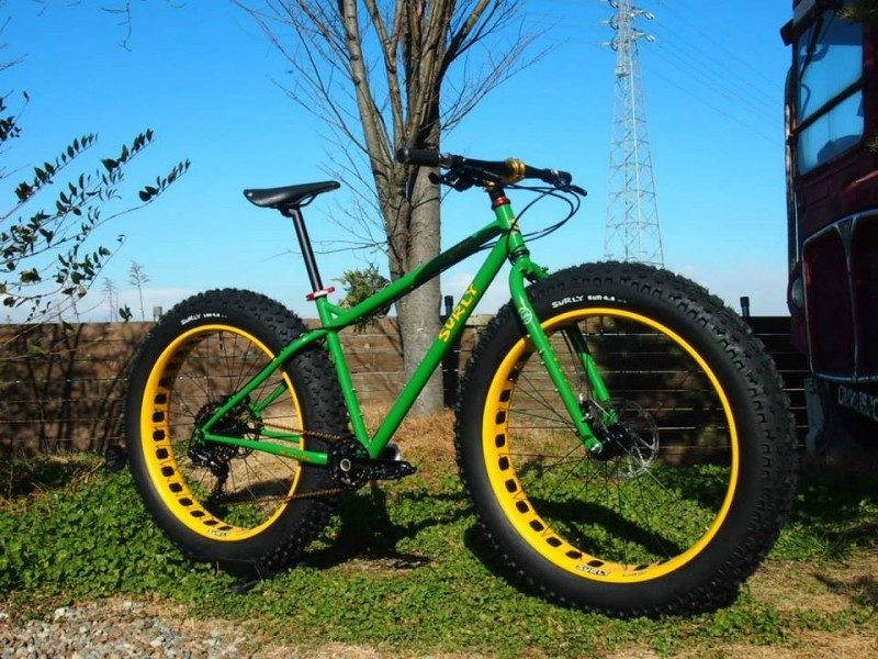 Right side view of a green Surly Moonlander fat bike on grass, in front of a double decker bus peeking out from a tree