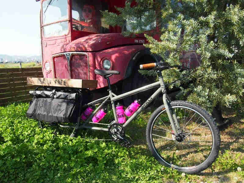 Right side view of a Surly Big Dummy bike, parked on grass in front of a double decker bus peeking out of the trees
