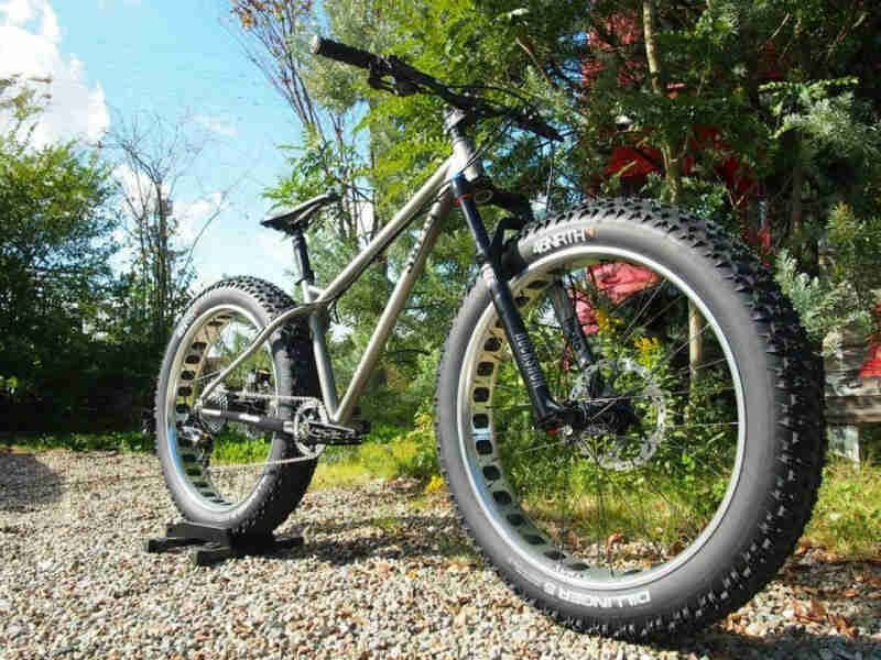 Right side view of a silver Surly fat bike, parked in a gravel patch with trees behind it