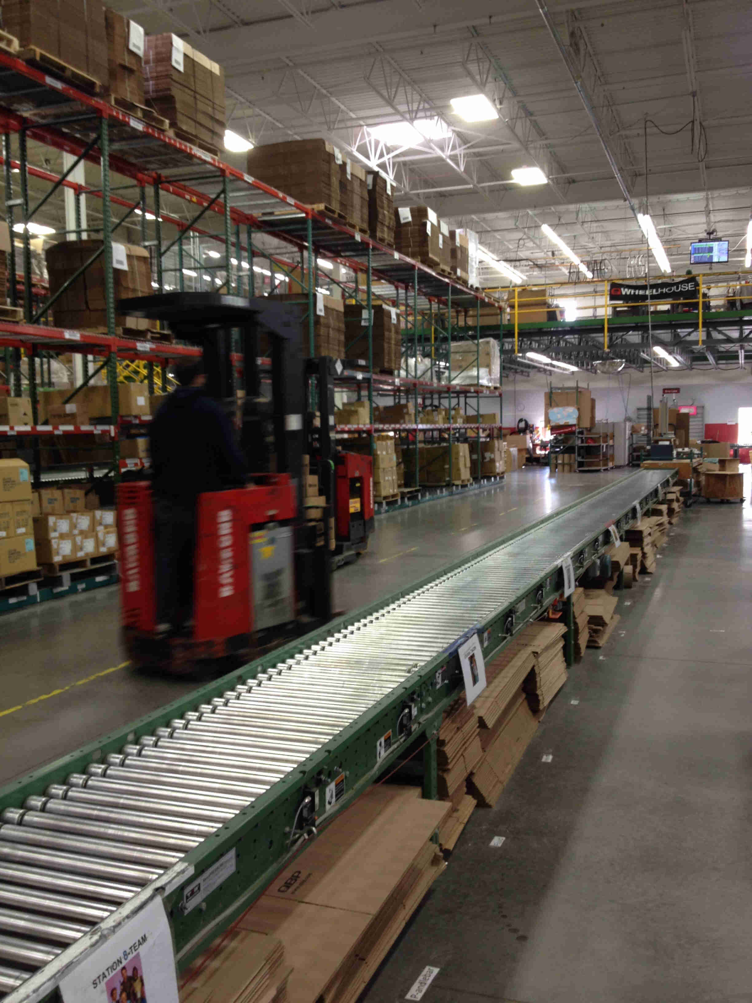 People on stand up forklifts, driving down a warehouse floor, alongside a conveyor line