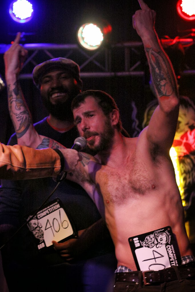 Front view of a shirtless person with a beard, on a stage with their middle fingers up, and person standing behind them