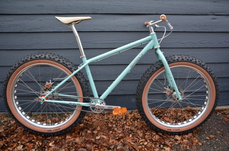 Right side view of a turquoise Surly fat bike, parked in leaves against a gray house wall 