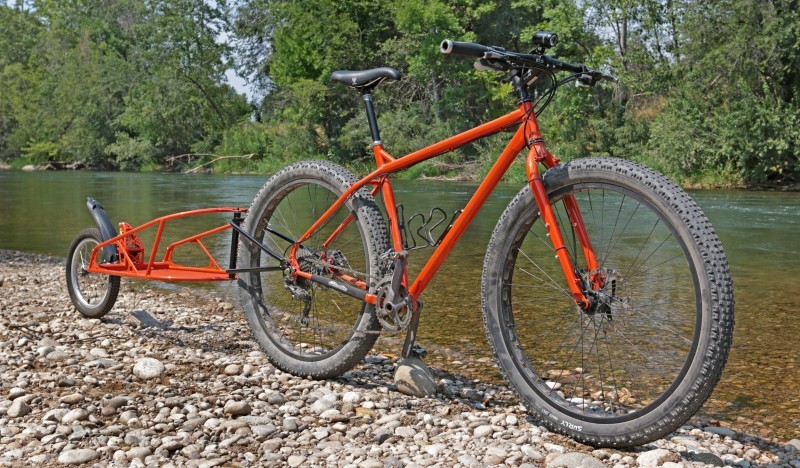 Right side view of a red Surly bike with a trailer, parked on a flat, rocky bank of a river with trees on the other side