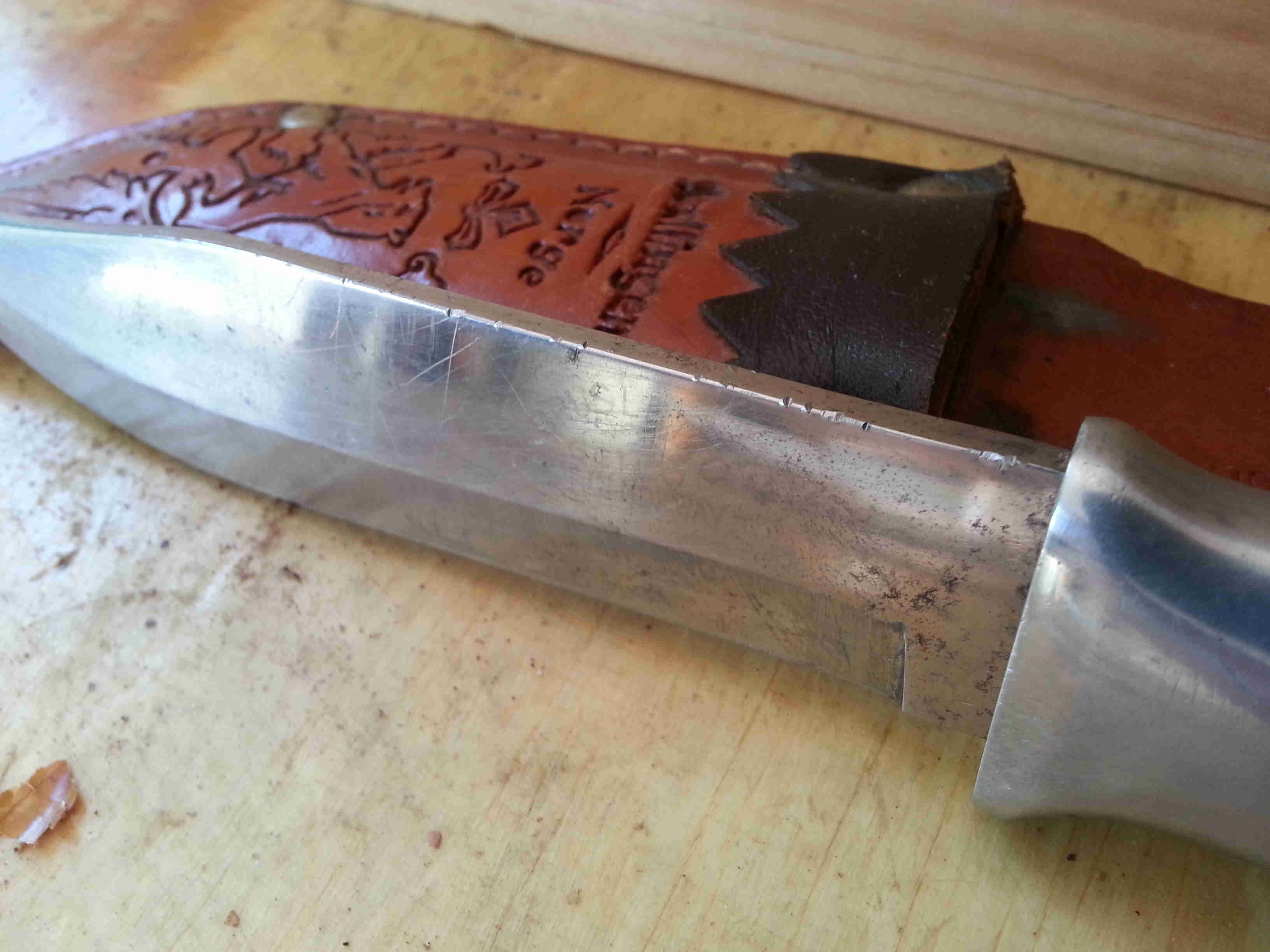 Horizontal view of a silver knife blade, standing on the cutting edge, leaning on a leather sheath