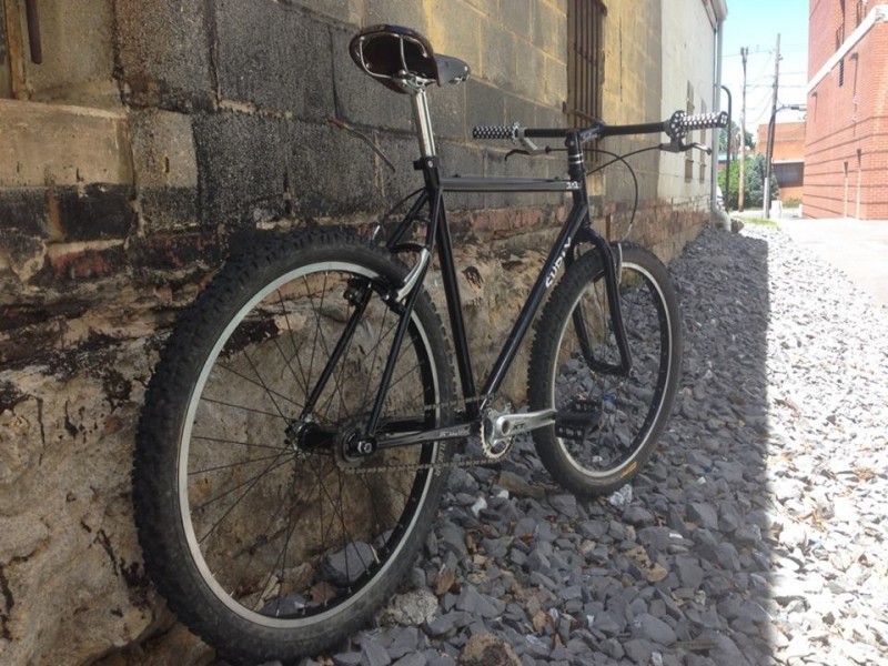 Rear, right side view of a black Surly 1x1 bike, leaning against a stone block wall, parked in a rock covered alley