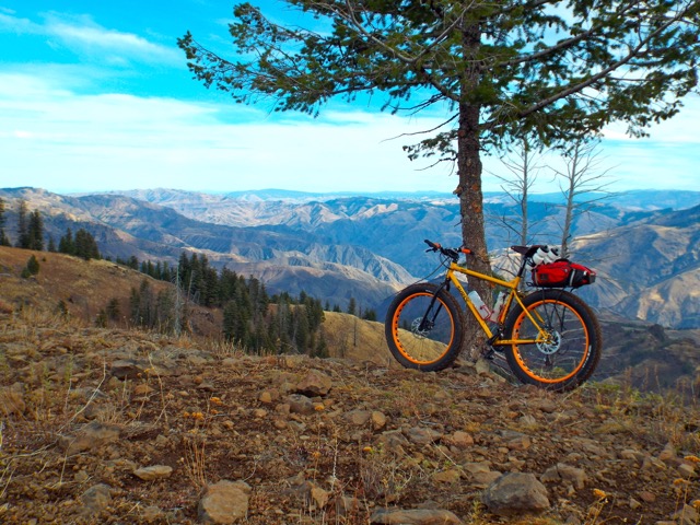 Left side view of a yellow Surly Pugsley fat bike, parked on a rocky hill against a tree, with a mountain range behind