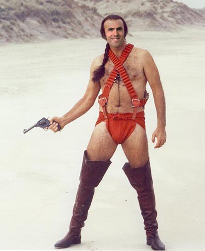 Front view of a person, wearing thigh high boots, shorts and suspenders, holding a gun, against a desert background